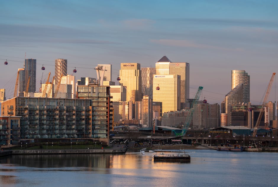 A panoramic view of the Canary Wharf financial district skyline at sunset, featuring tall modern office buildings with glass facades reflecting the orange and pink hues of the sky. In the foreground,