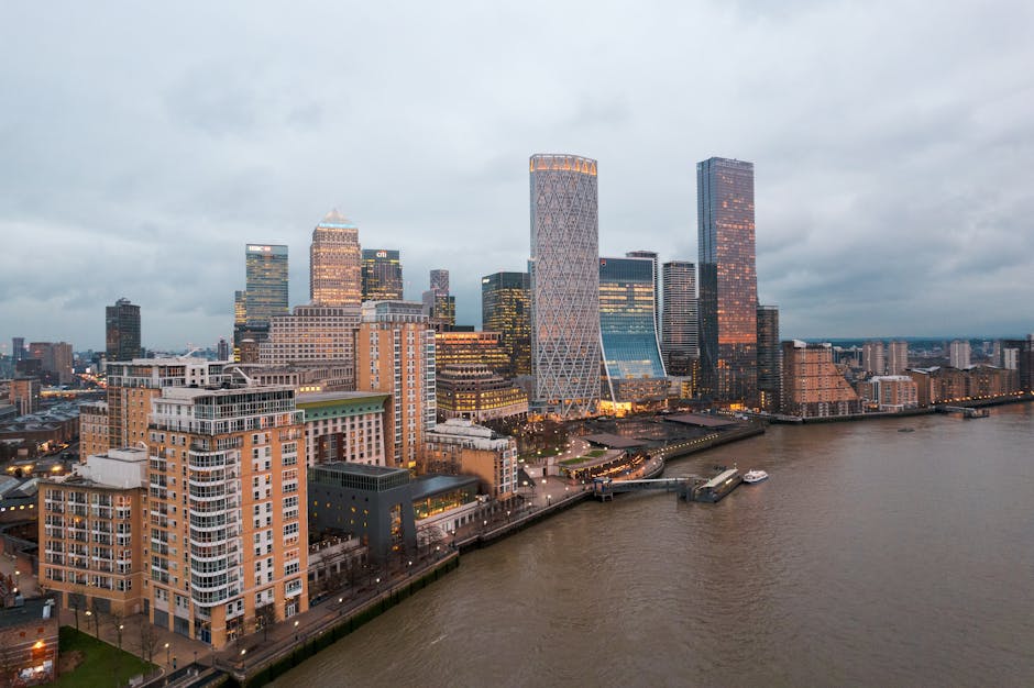 A panoramic view of the Canary Wharf financial district skyline at sunset, featuring tall modern office buildings with glass facades reflecting the orange and pink hues of the sky. In the foreground,